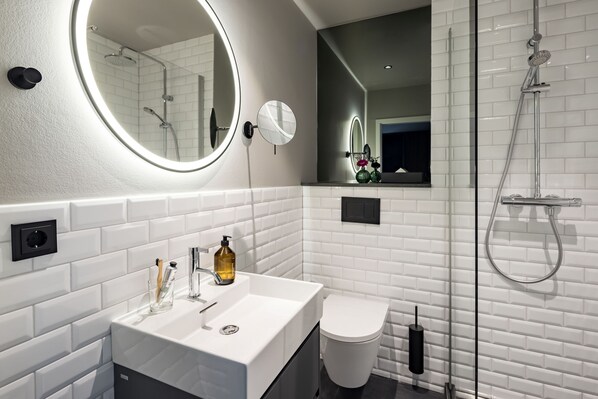 Une salle de bain moderne avec un mur recouvert de carreaux de métro blancs, un grand miroir rond avec éclairage intégré, un lavabo blanc et une douche équipée d’un pommeau de douche à main.