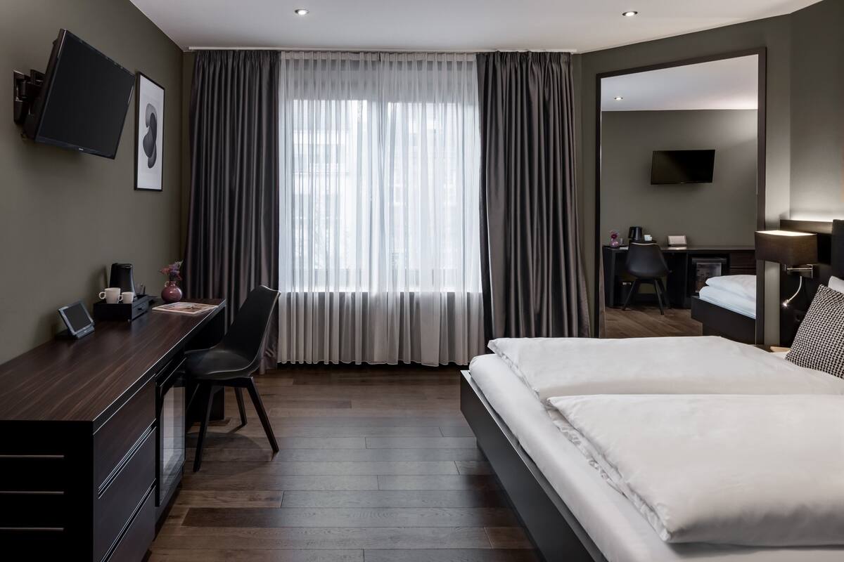 A hotel room with a bed, a desk with a chair, a TV mounted on the wall, and a large window with curtains.