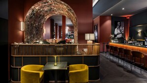 Uma área de bar com um arco de madeira curvado, duas cadeiras amarelas, uma mesa e um balcão com bancos.