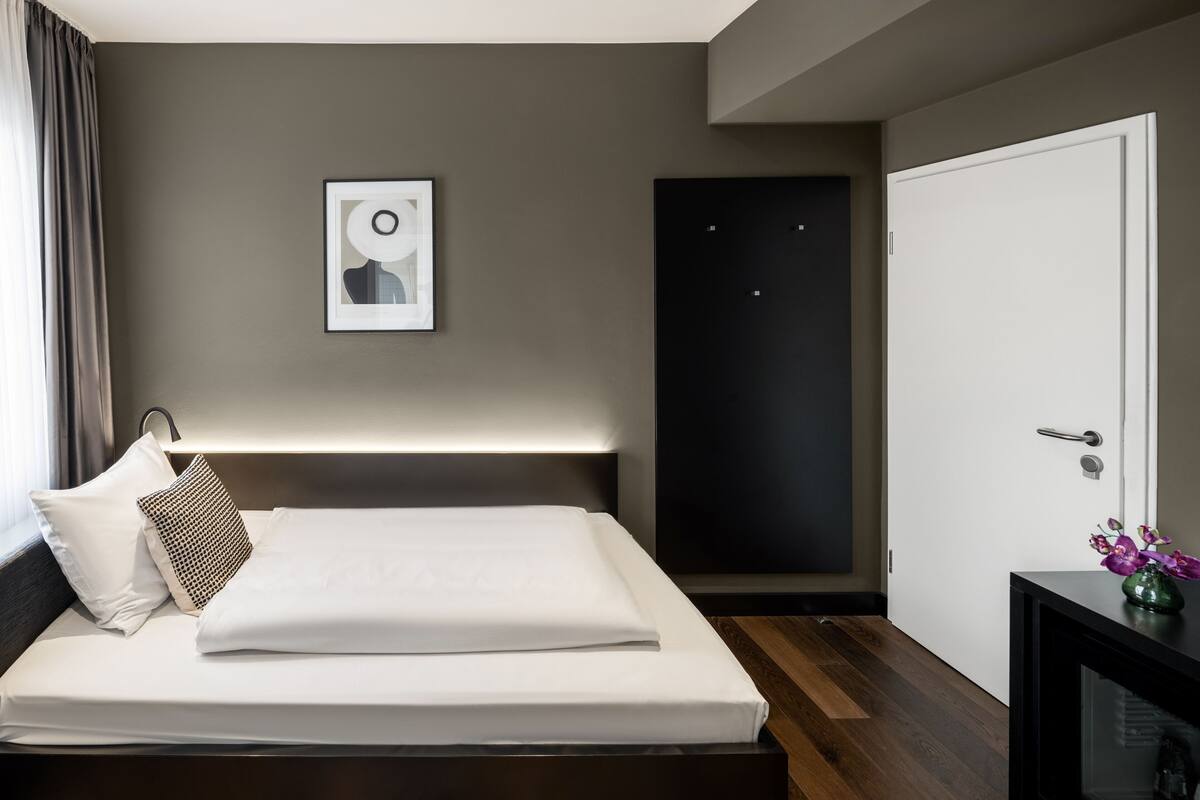 A modern hotel room with a neatly made bed, a framed picture on the wall, a dark door, and a small table with a vase of flowers.