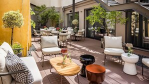 Uma área de estar ao ar livre com vários conjuntos de assentos, incluindo sofás e cadeiras, e diversos elementos decorativos como plantas e mesas.