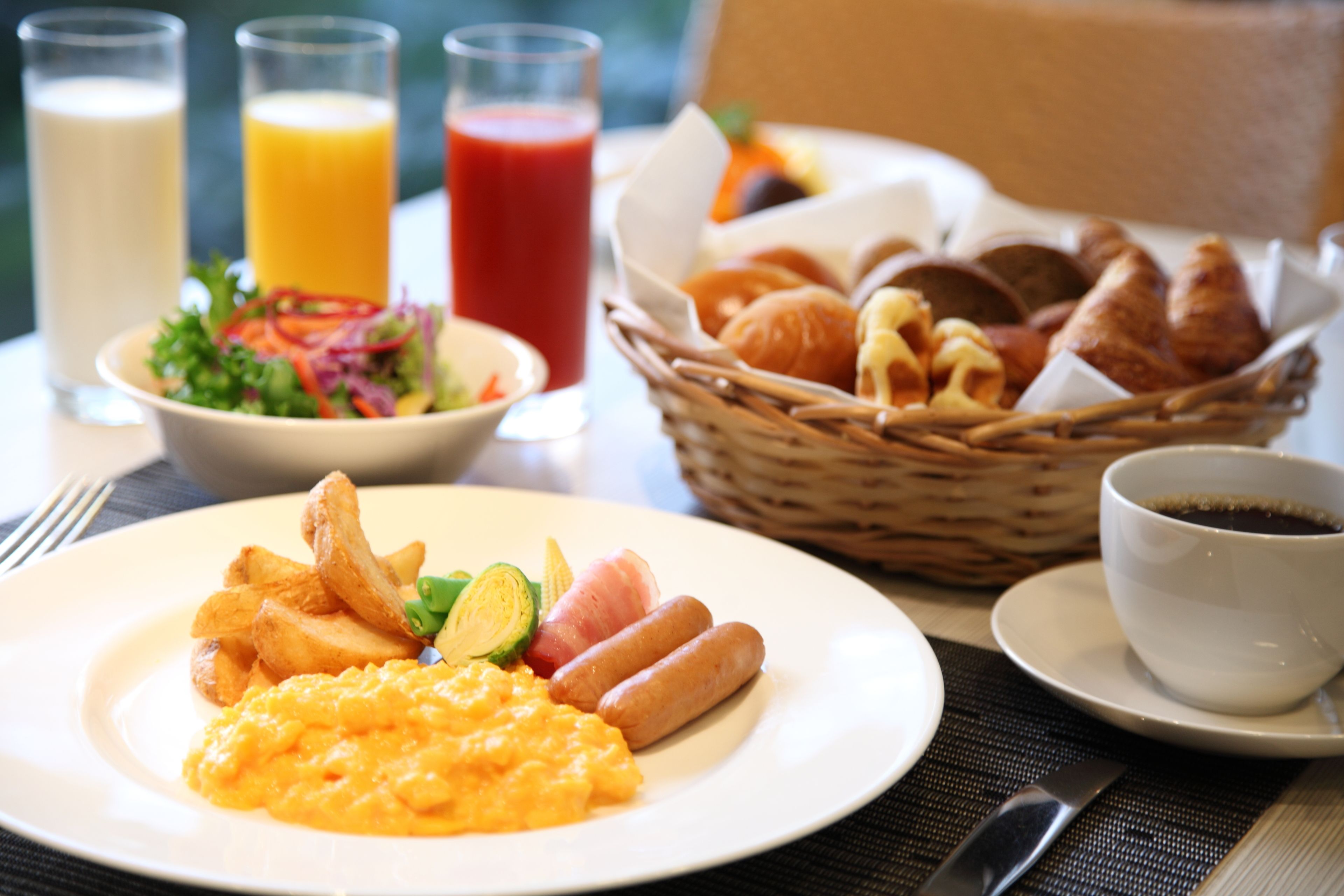 Une table dressée pour le petit-déjeuner avec des œufs brouillés, des saucisses et des frites dans une assiette, un saladier rempli de salade et un panier de croissants. On y trouve également des verres de jus d’orange et un verre de jus rouge, ainsi qu’une tasse de café.