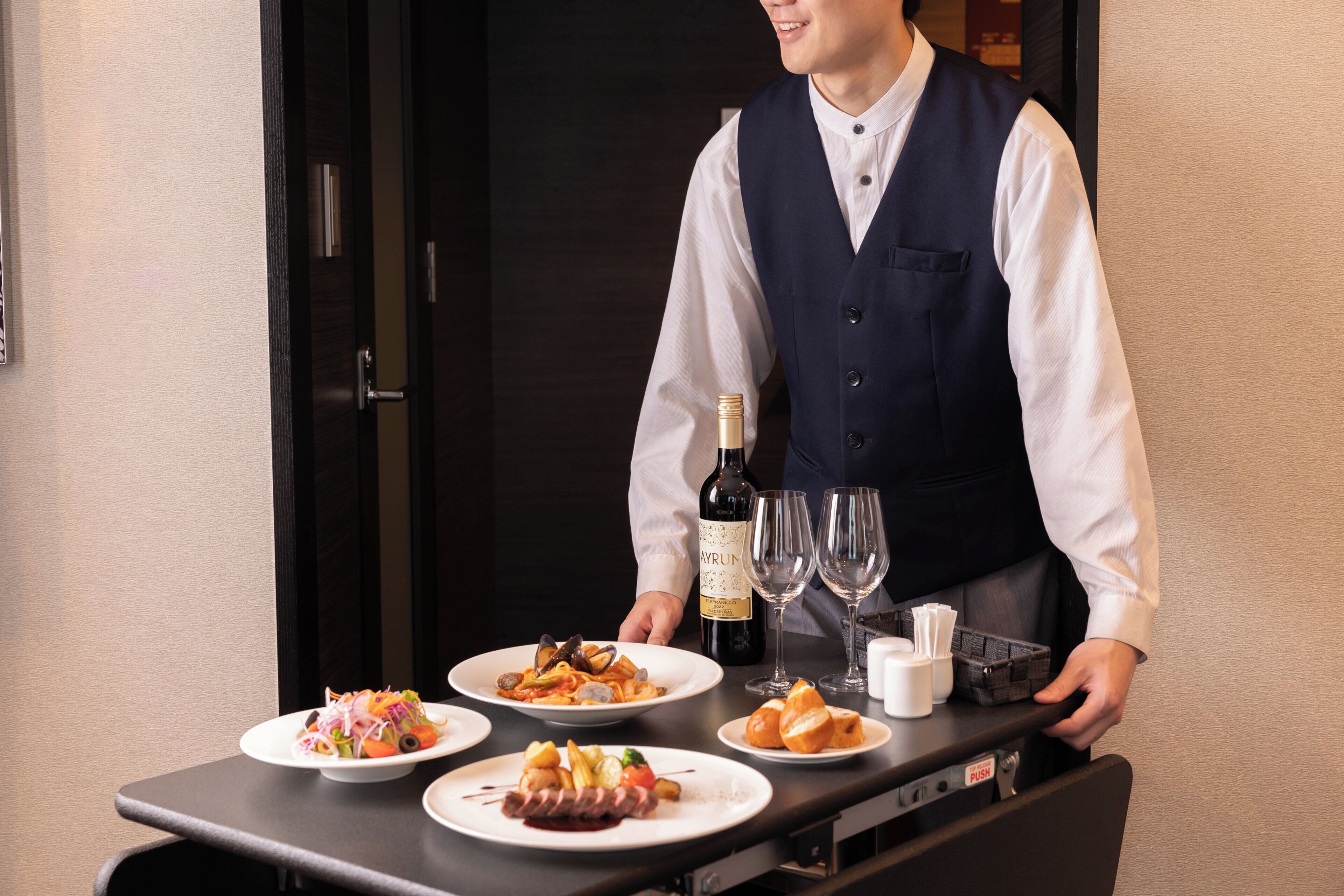 A hotel room with a dining table set with plates of food, a bottle of wine, and glasses.