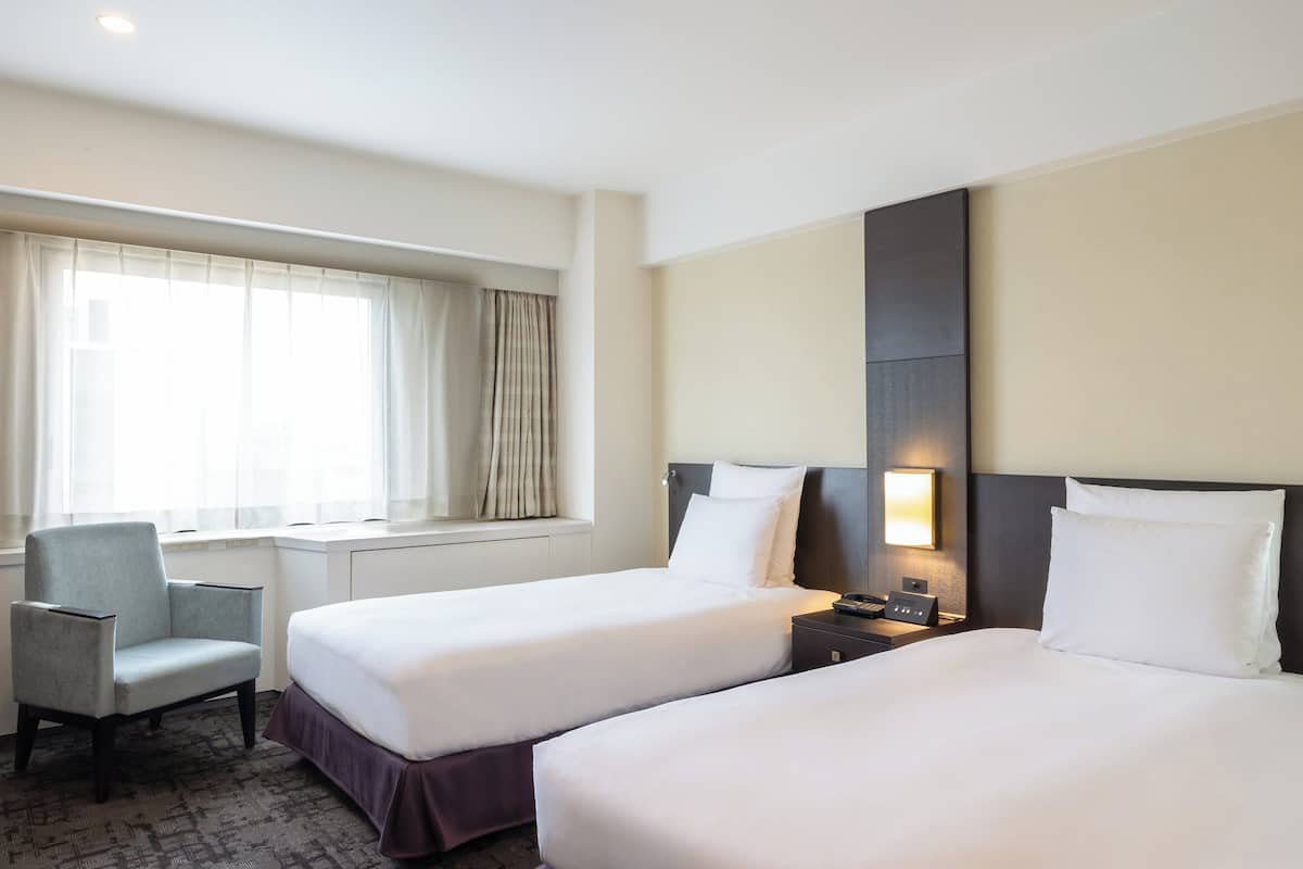 Une chambre d’hôtel avec deux lits, une chaise, une table de chevet et une fenêtre avec des rideaux.