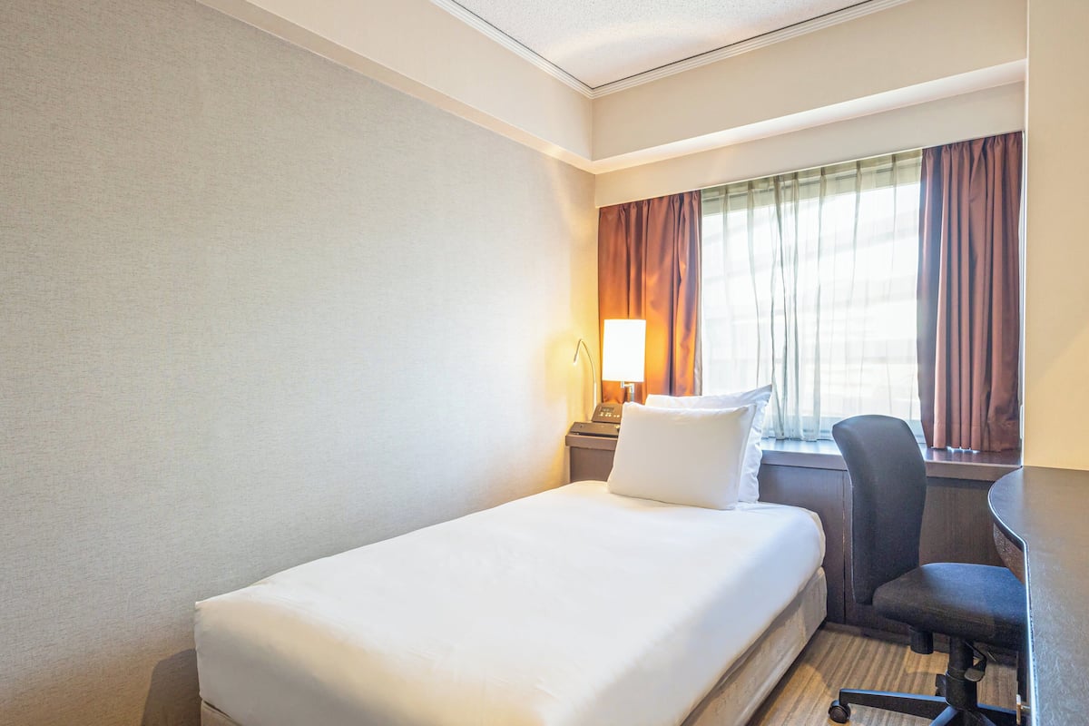 Une chambre d’hôtel avec un lit simple, un bureau avec une chaise, une lampe et une fenêtre avec des rideaux.