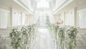 Um salão amplo com teto alto e lustres, decorado com arranjos florais e uma cruz no extremo.