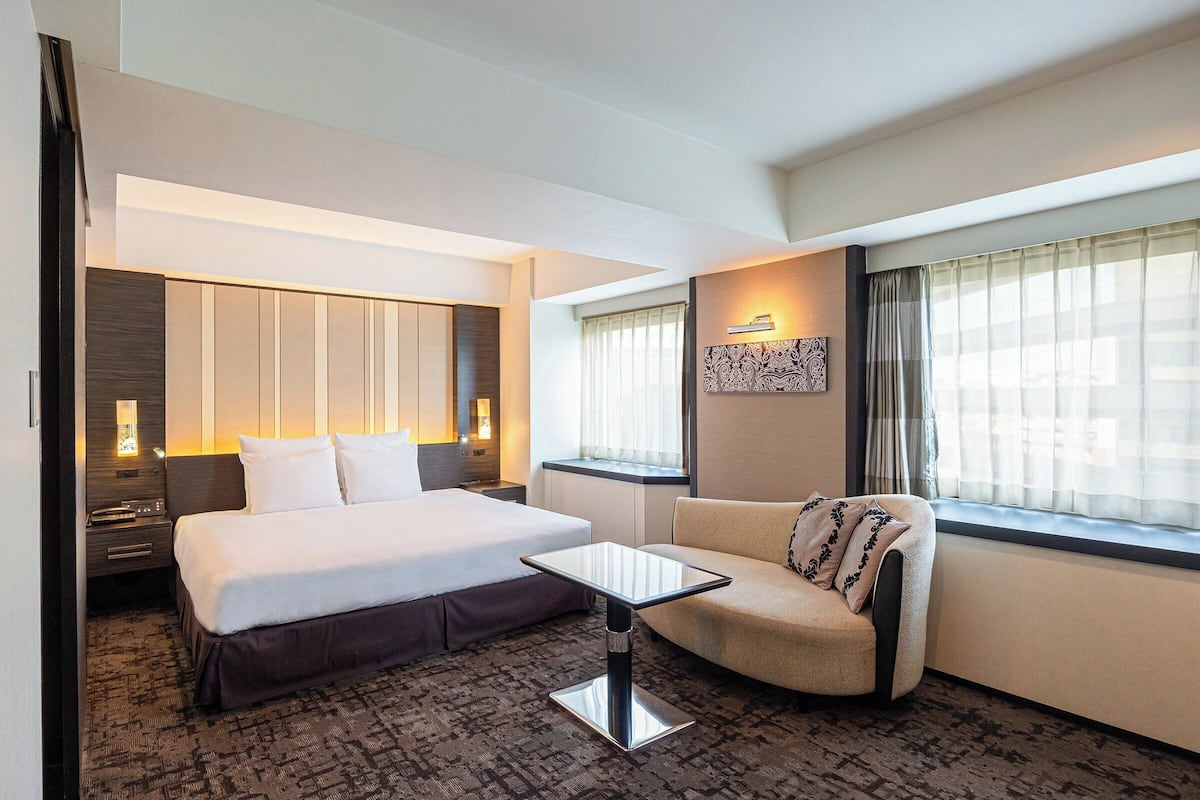 Une chambre d’hôtel avec un lit, un canapé, une petite table, une table de chevet et une fenêtre avec des rideaux.
