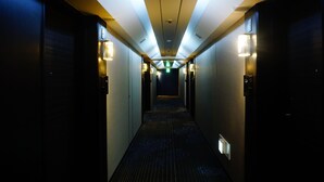 Corredor longo e estreito de um hotel, com iluminação no teto e placas de saída de emergência.