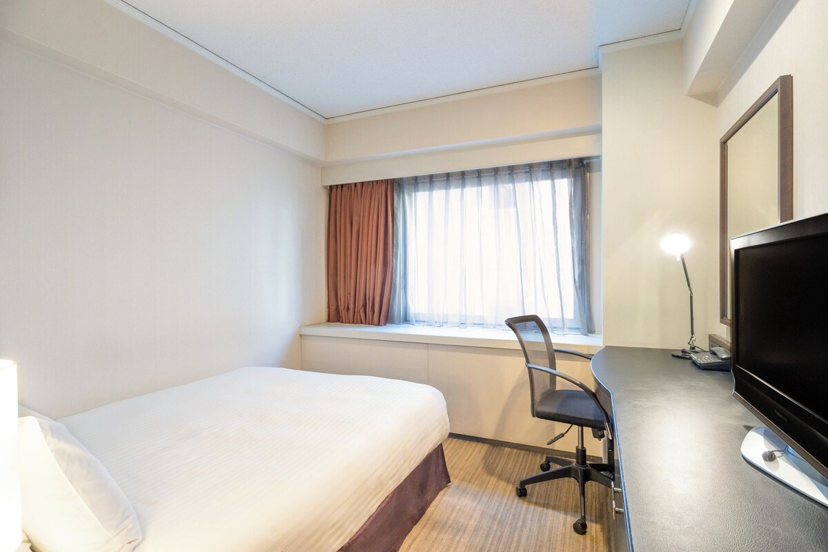 A hotel room with a bed, a desk with a computer, a TV, and a window with curtains.