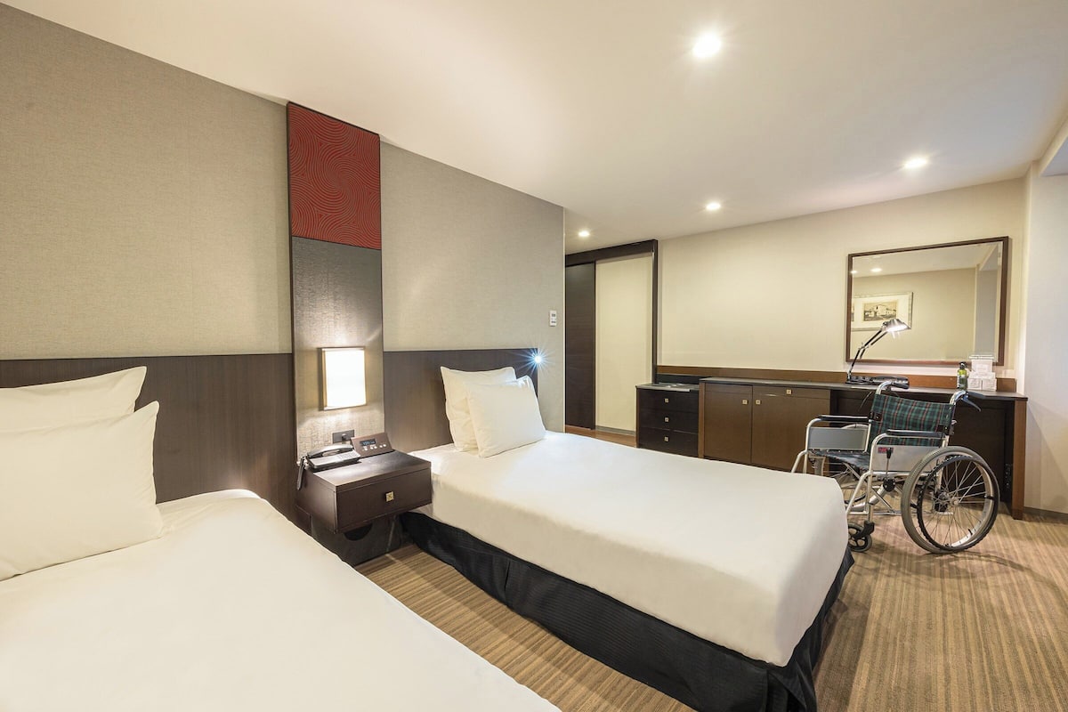 Une chambre d’hôtel avec deux lits, un fauteuil roulant, un bureau et un miroir.