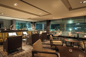 A modern hotel lobby with seating areas, a bar, and large windows offering city views.