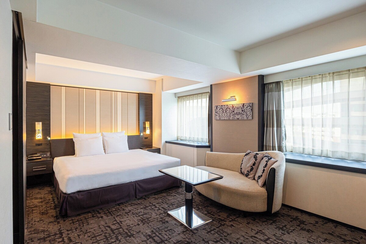 A hotel room with a bed, a sofa, a small table, a nightstand, and a window with curtains.