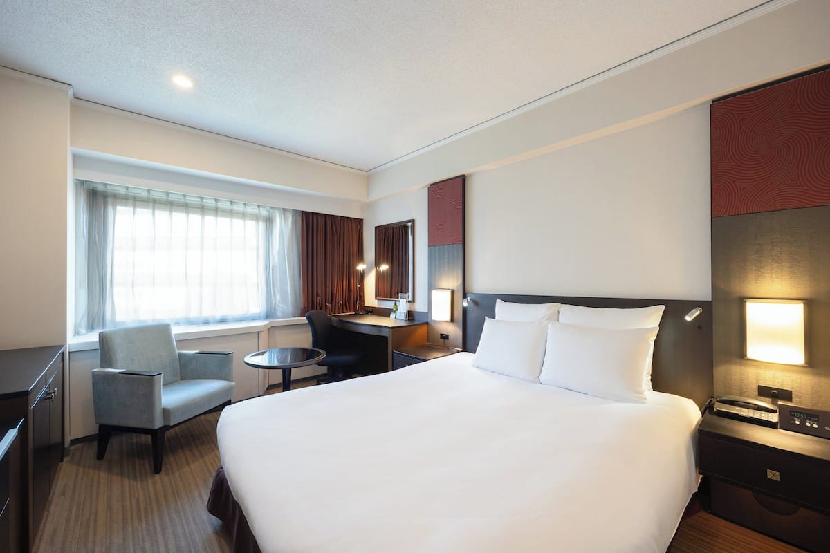 Une chambre d’hôtel avec un grand lit, un bureau, une chaise, une petite table, une lampe et une fenêtre avec des rideaux.