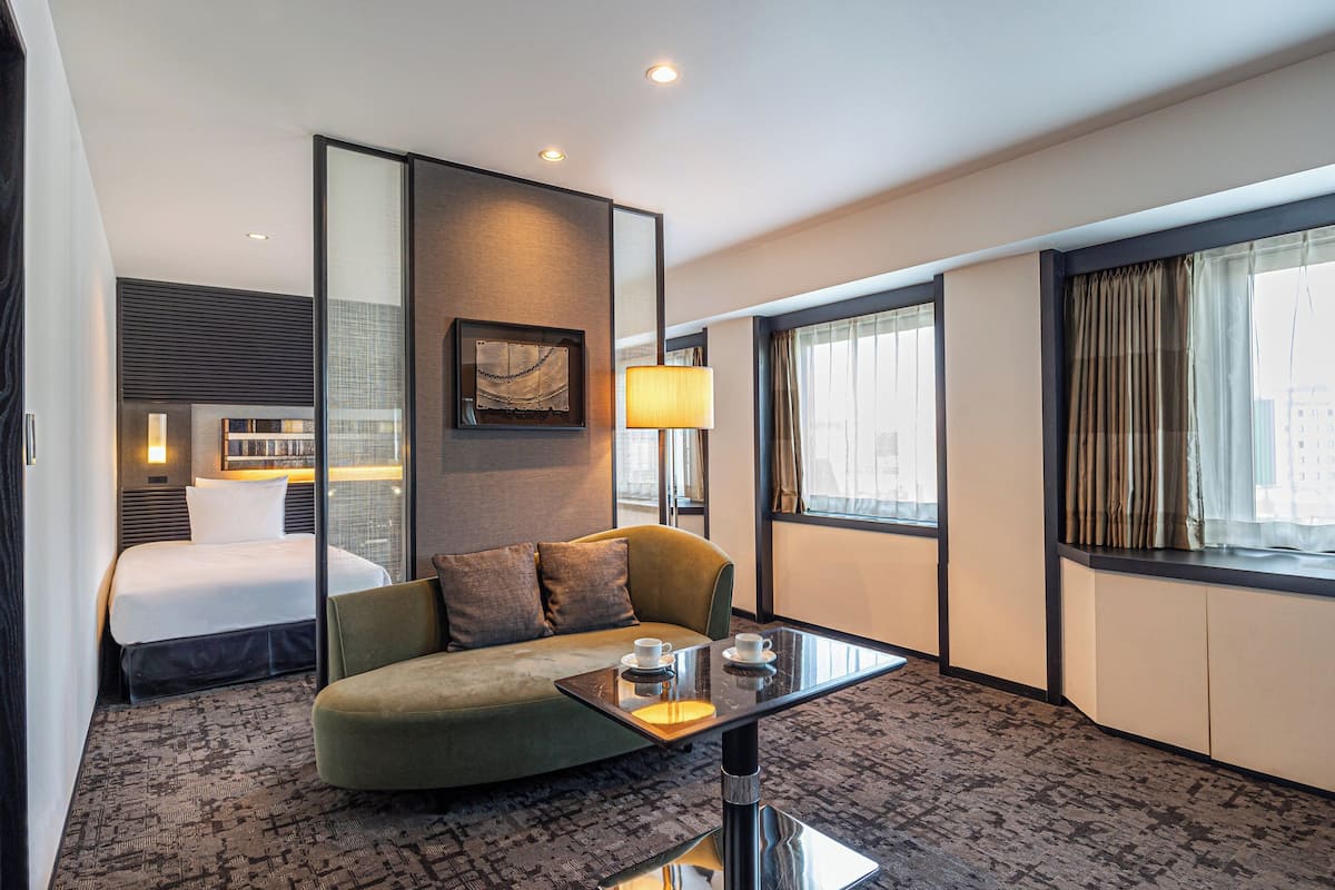 Une chambre d’hôtel avec un lit, un canapé, une table en verre avec deux tasses, une lampe et de grandes fenêtres avec des rideaux.