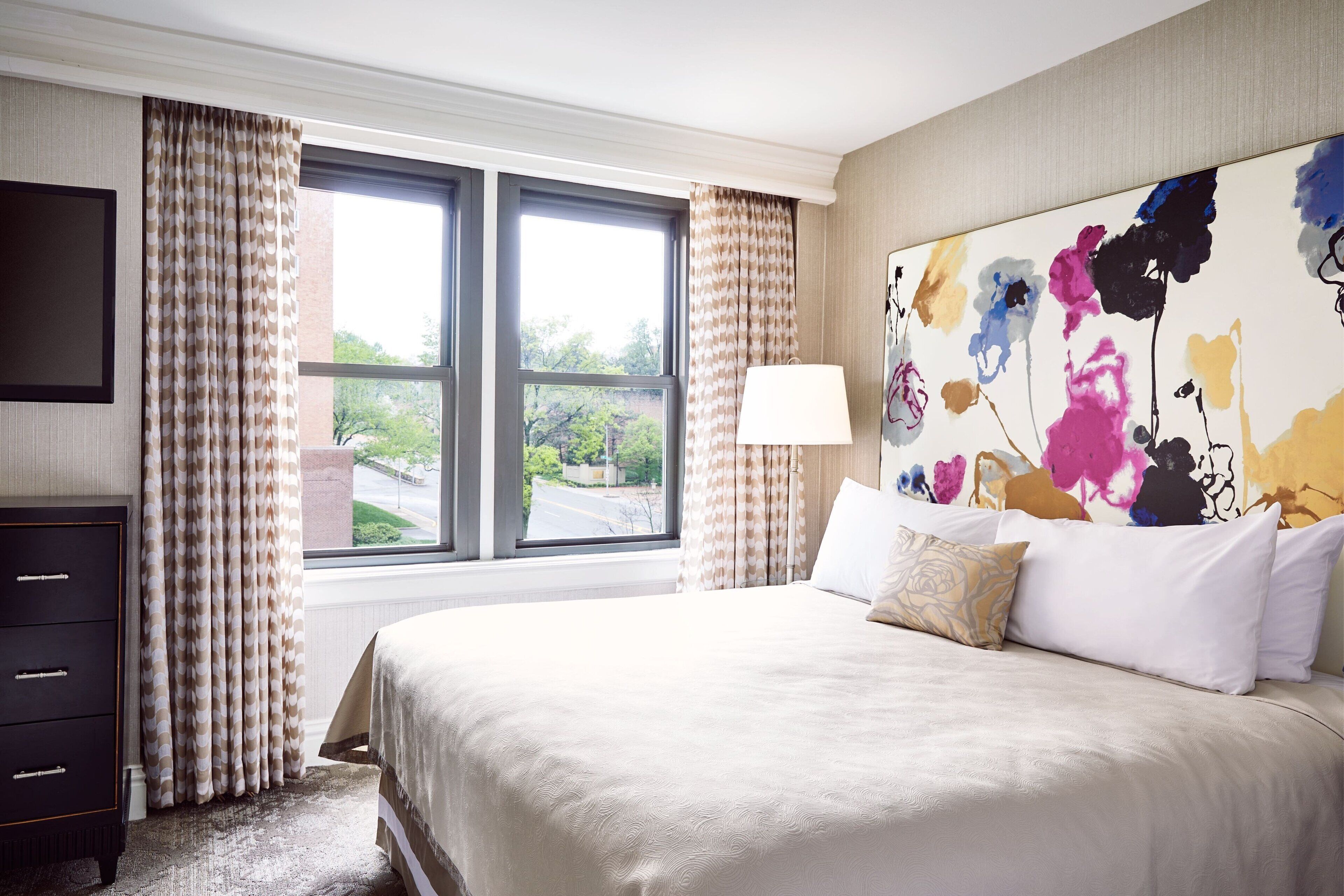A hotel room with a large bed, a flat-screen TV, a window with curtains, and a colorful abstract painting on the wall.