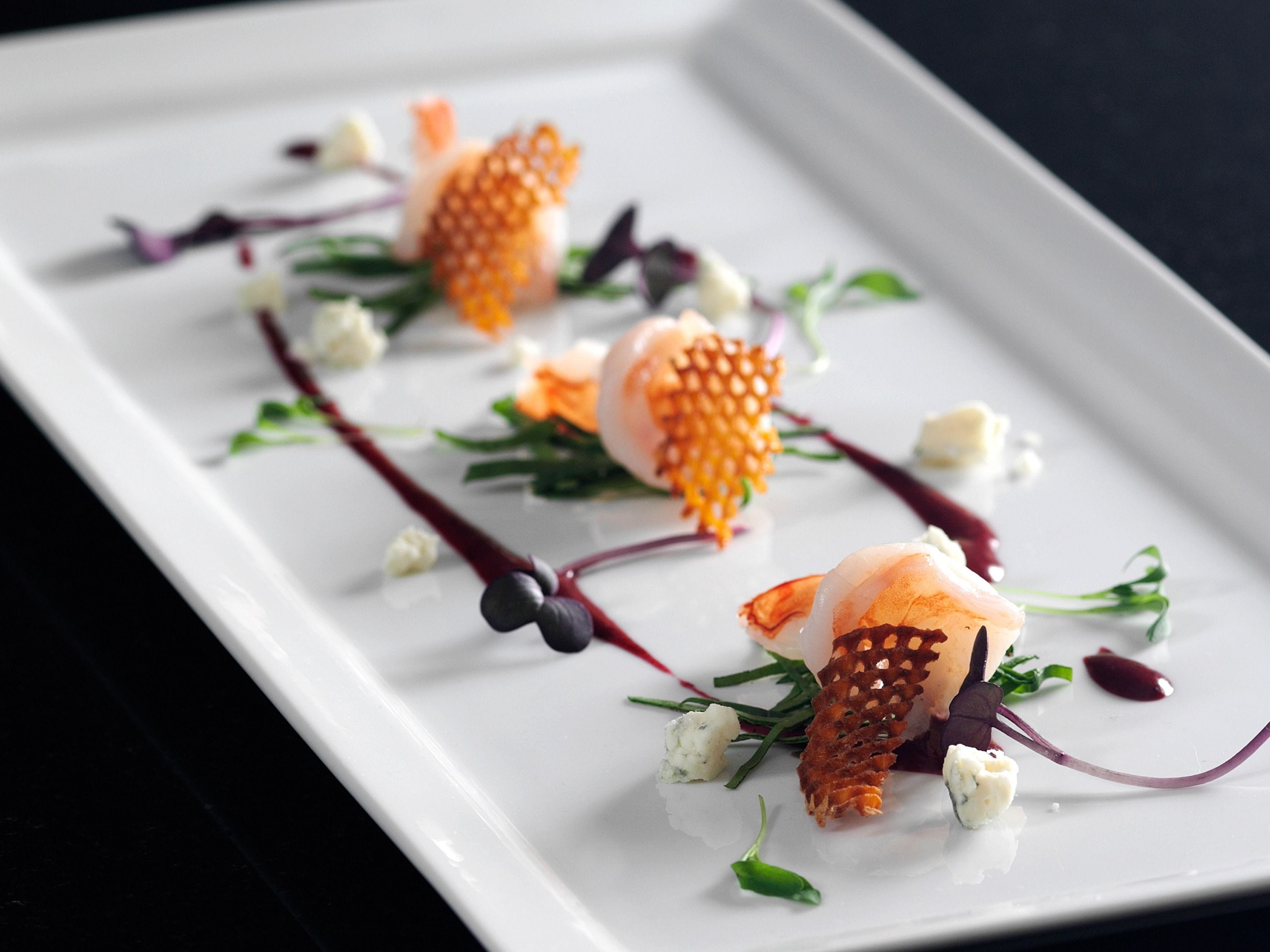 Un piatto bianco rettangolare con un piatto di pesce, guarnito con erbe aromatiche verdi e elementi viola, su uno sfondo scuro.