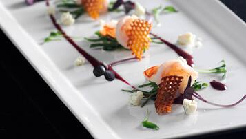 Un piatto bianco rettangolare con un piatto di pesce, guarnito con erbe aromatiche verdi e elementi viola, su uno sfondo scuro.