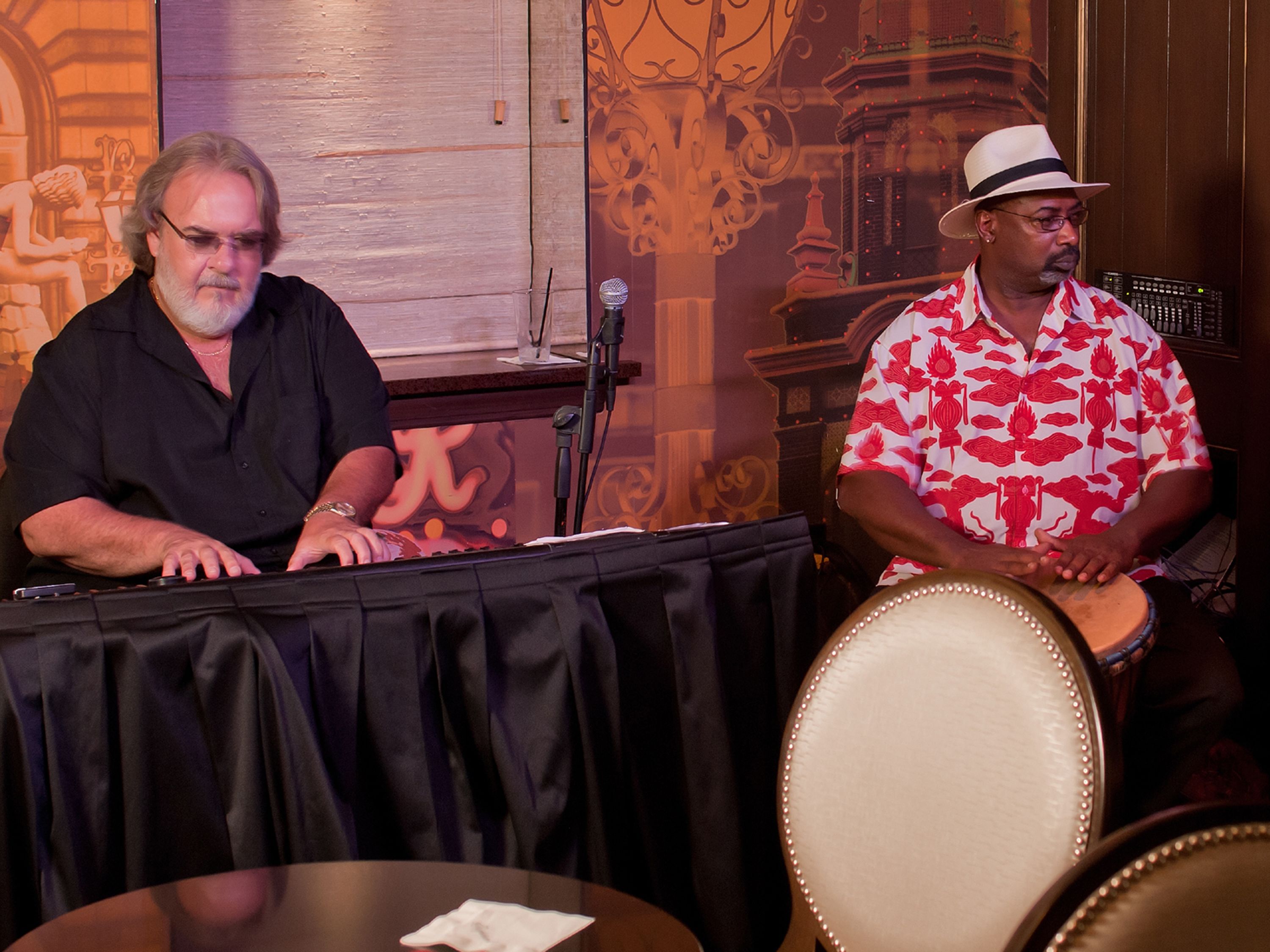 Two individuals seated at a table with a microphone stand, a keyboard, and a drum.