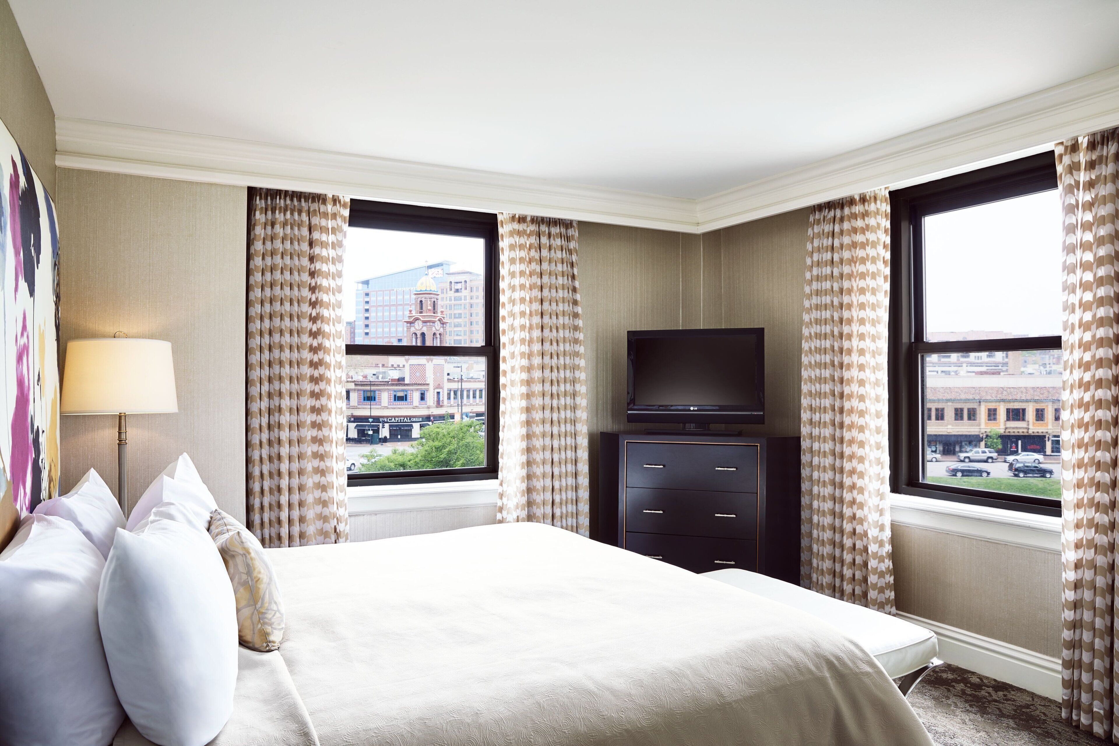 Una habitación de hotel con cama, televisor, cómoda y ventanas grandes con cortinas.
