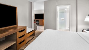A modern hotel room with a large bed, wooden furniture, a flat-screen TV, a small kitchenette, and a glass door leading to a bathroom.
