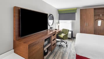 Ett hotellrum med en stor platt-TV, ett skrivbord med en stol, en säng, en garderob och ett fönster med utsikt.