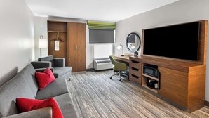A modern hotel room with a grey sofa, red cushions, a desk with a computer, a flat-screen TV, and a wooden cabinet.