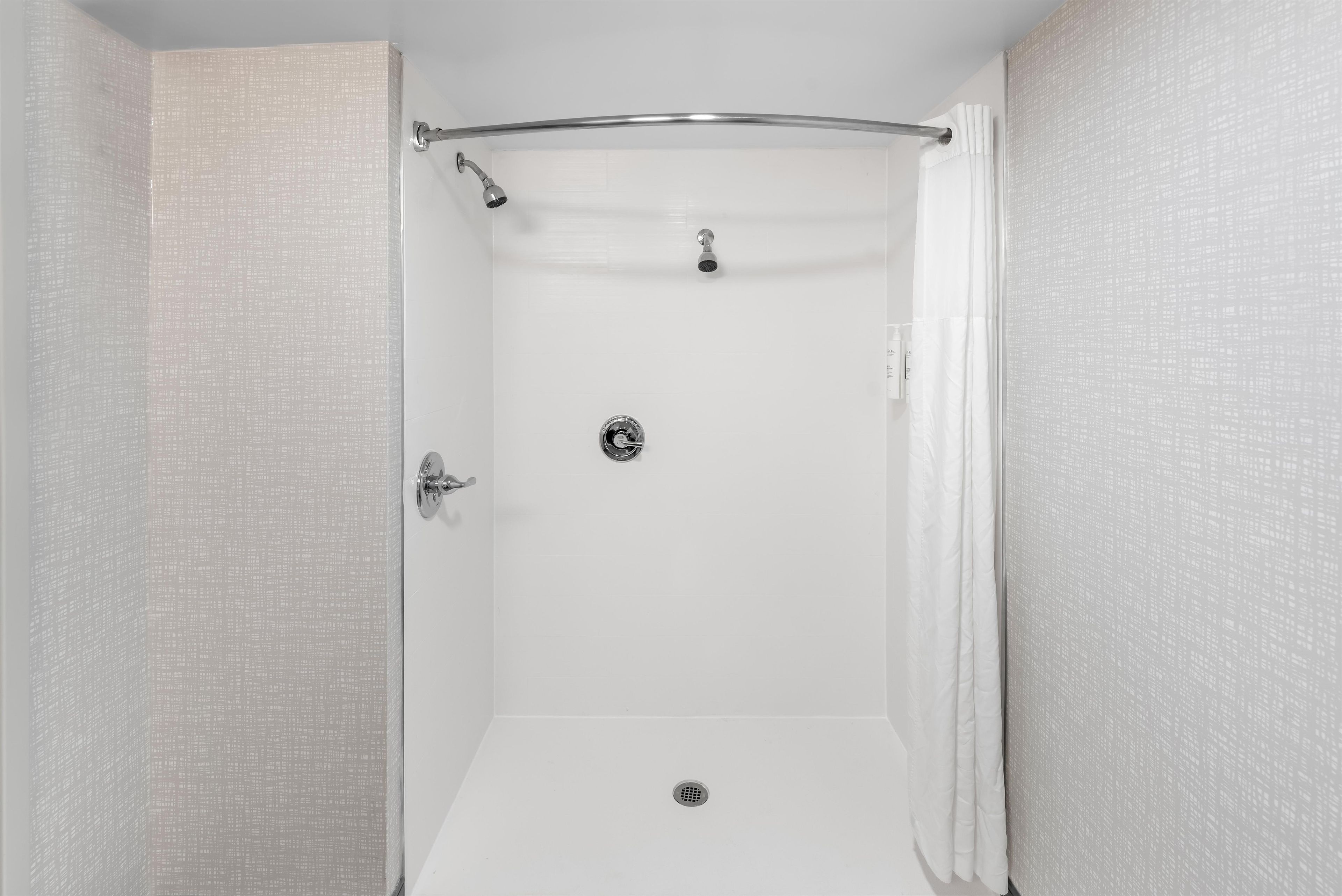 A walk-in shower with a glass door, a showerhead, a handheld showerhead, a control panel, and a drain.