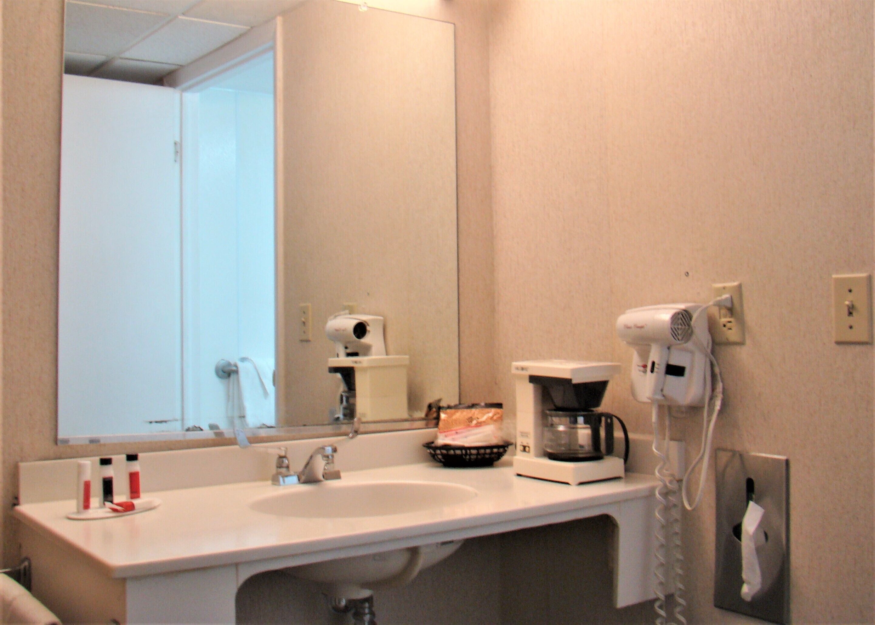 Un baño con lavamanos blanco, espejo, secador de pelo y cafetera.
