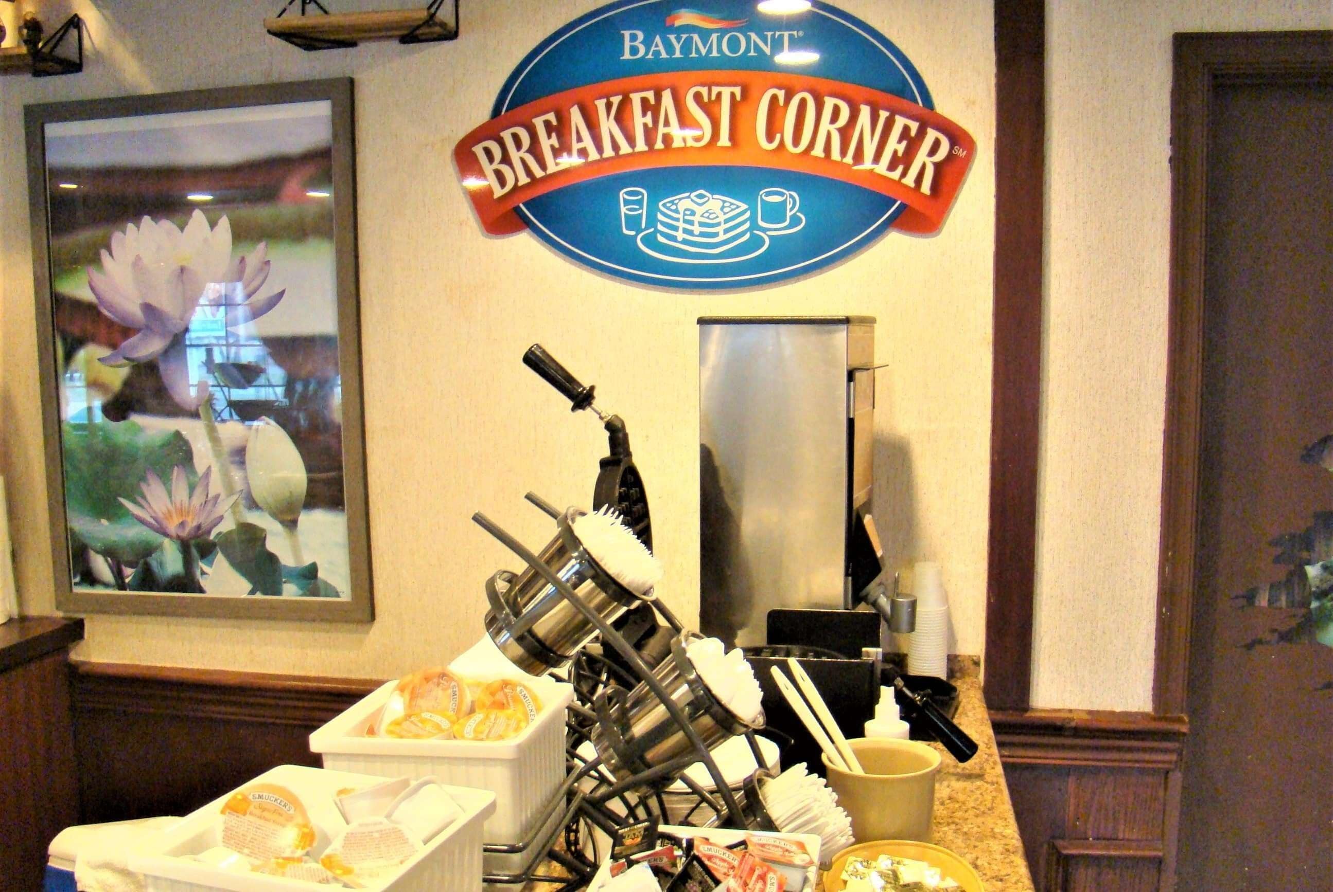 A breakfast corner with a sign reading "Baymont Breakfast Corner," a coffee machine, and a dish rack with utensils.