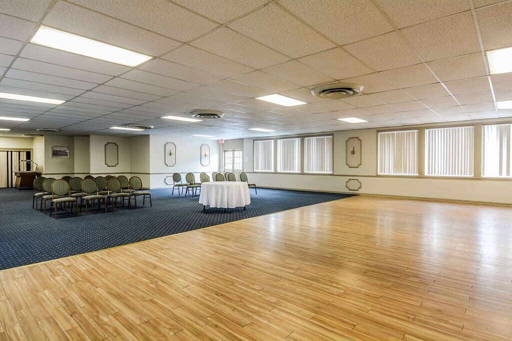 A large, well-lit conference room with a wooden floor, multiple tables with chairs, and large windows providing natural light.