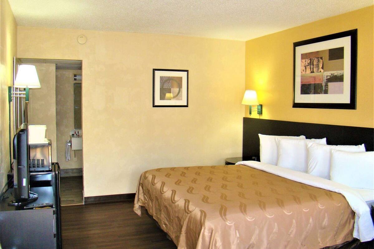A hotel room with a bed, a desk with a lamp, a coffee machine, a framed picture on the wall, and a door leading to a bathroom.