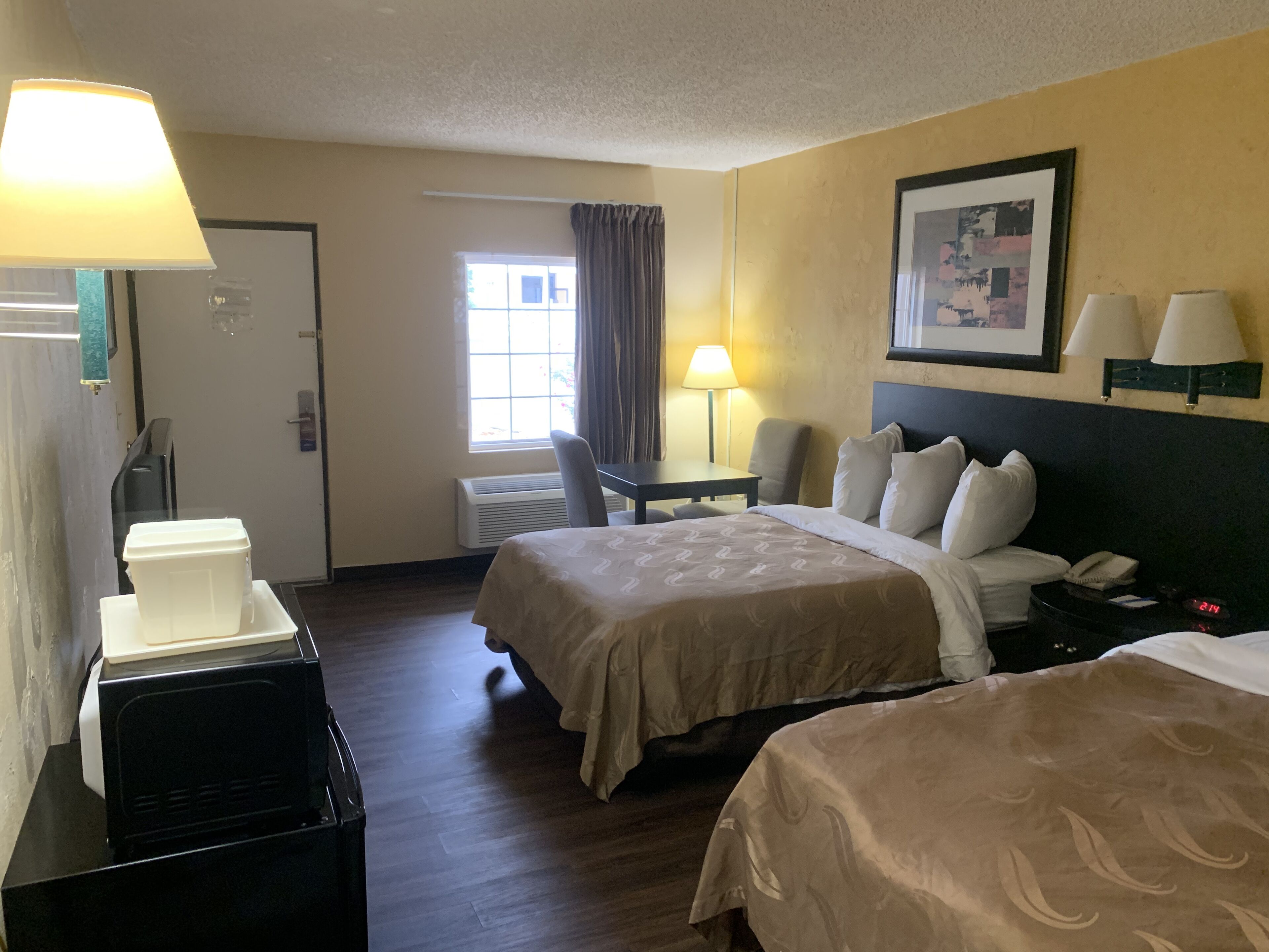 Habitación de hotel con dos camas, un escritorio, una silla, una lámpara, un calefactor y un cuadro en la pared.