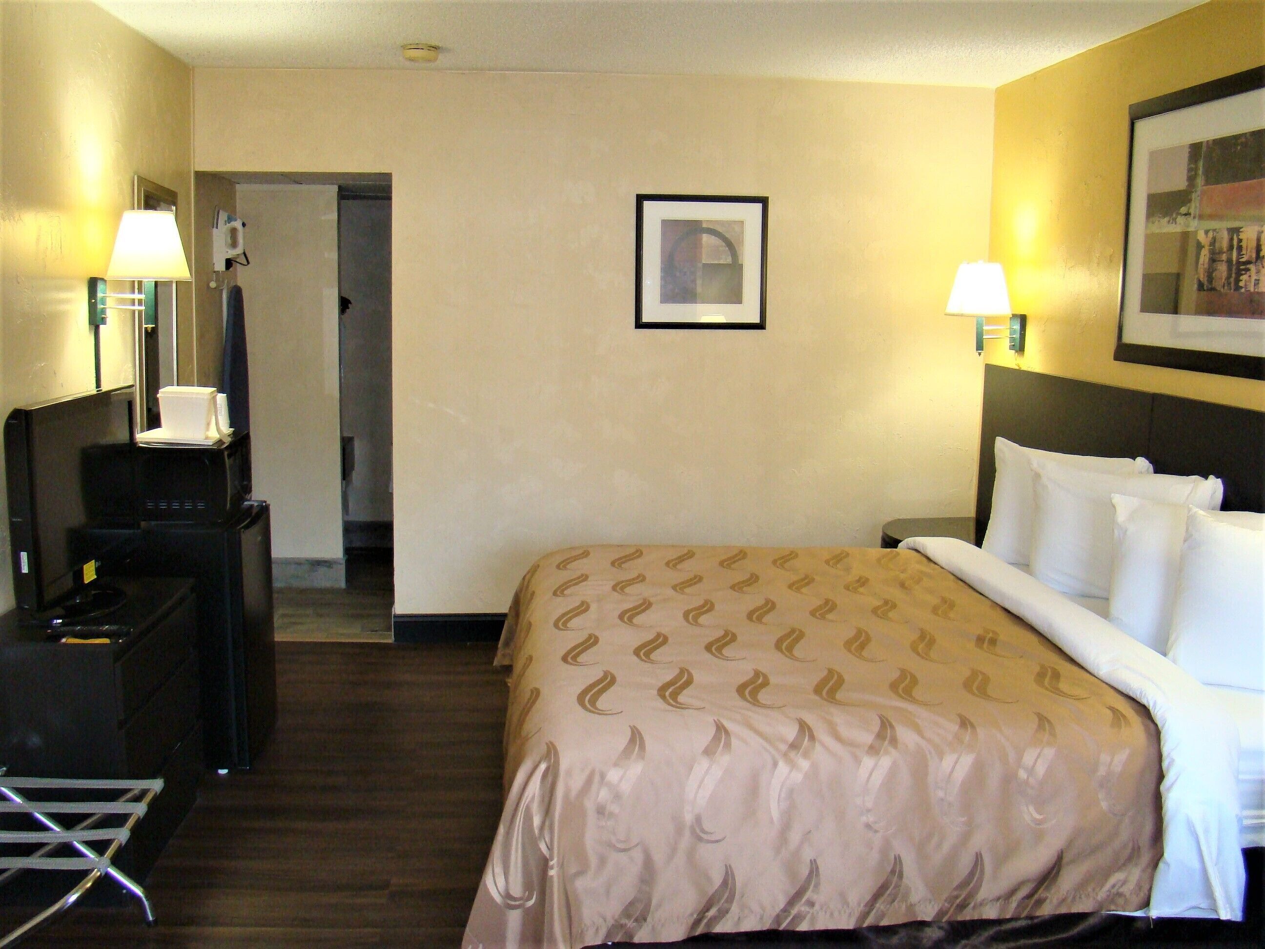 A hotel room with a bed, bedside lamps, a desk with a lamp, a framed picture on the wall, and a door leading to another room.