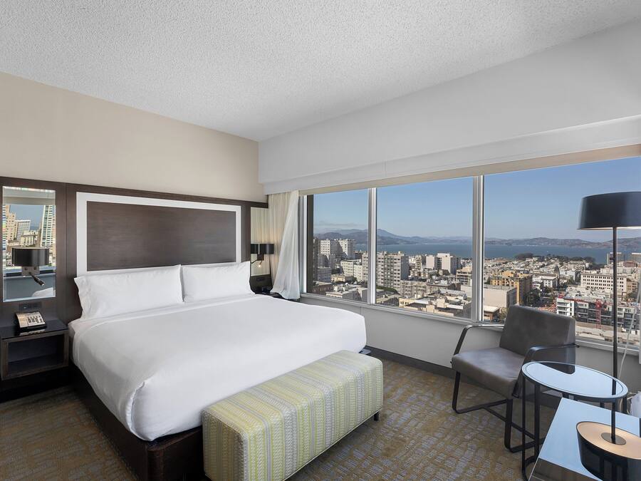 A hotel room with a large bed, a small table, a chair, and a view of the city through large windows.