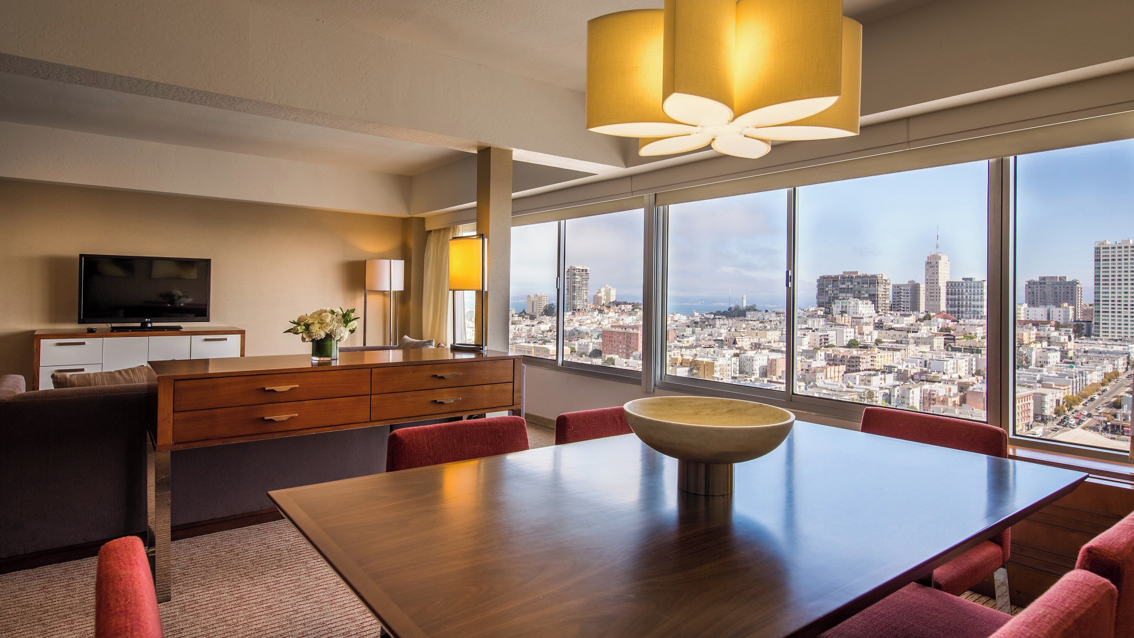 Une chambre d’hôtel moderne offrant une vue imprenable sur la ville, avec une table à manger en bois et un téléviseur posé sur la commode.