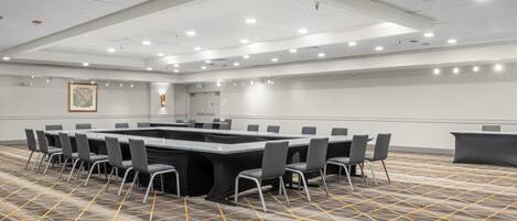 Un salón de conferencias con una gran mesa rectangular rodeada de sillas, una alfombra estampada y un techo blanco con iluminación empotrada.