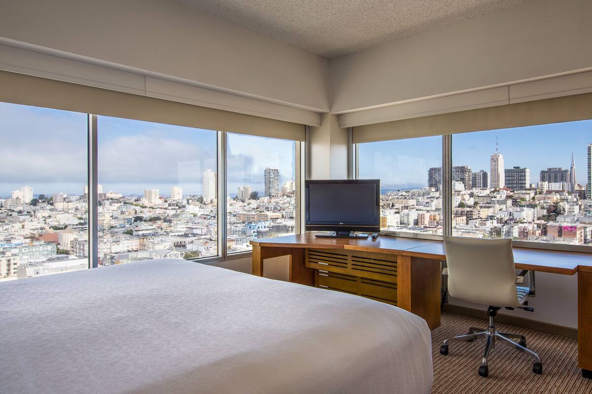 A hotel room with a large bed, a desk with a TV, and a view of a cityscape through large windows.