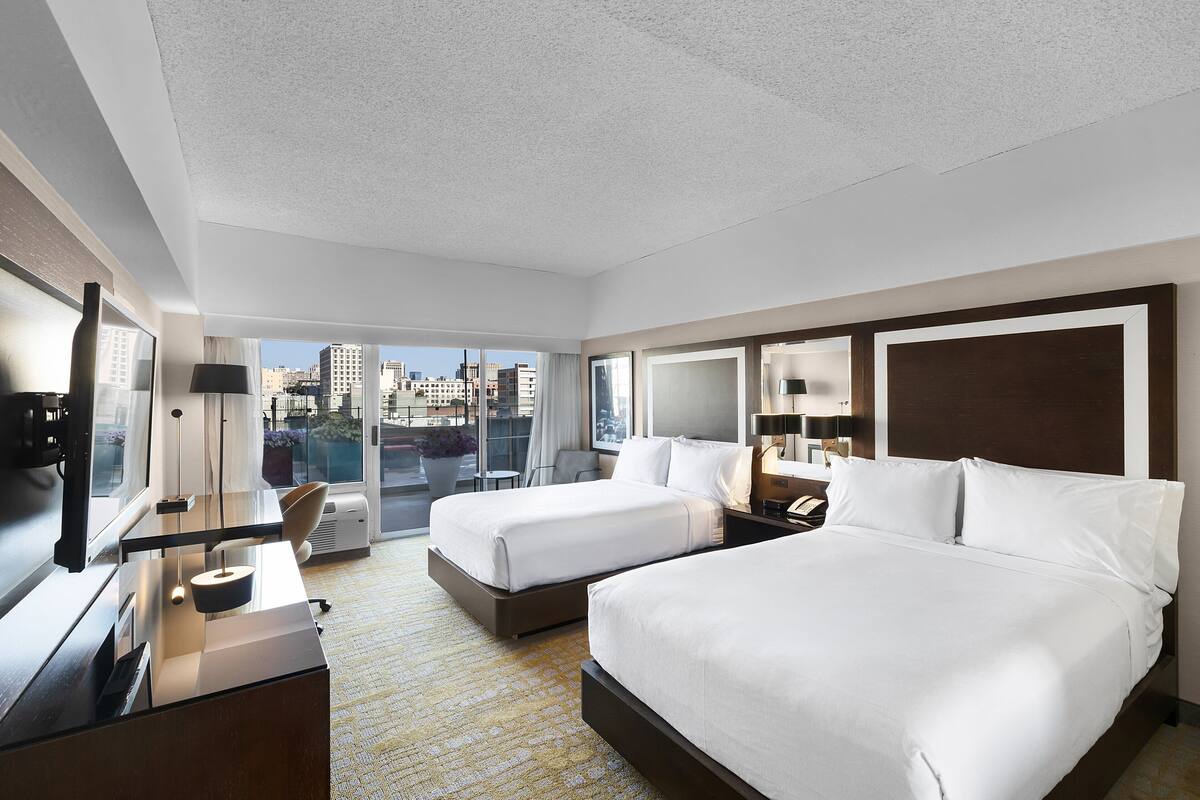 A hotel room with two beds, a desk, a chair, and a large window offering a city view.