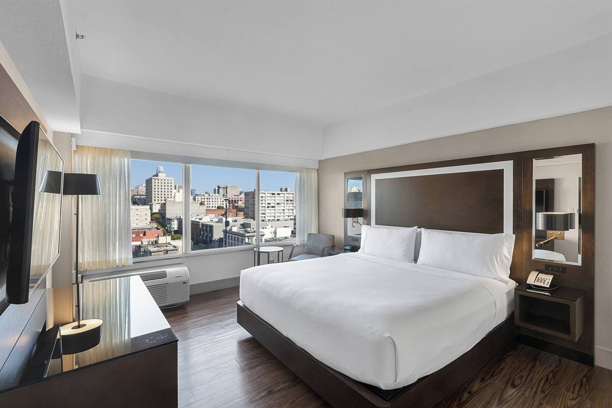 A hotel room with a large bed, a TV, a desk, a chair, a small table, a nightstand, and a city view.