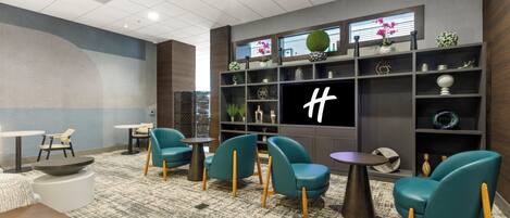 Um hall de hotel moderno com cadeiras verde-água, uma televisão grande e uma parede decorativa com prateleiras.