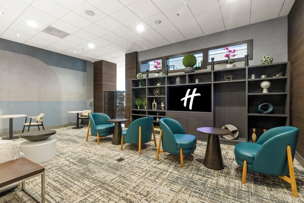 En modern hotellobby med turkosa fåtöljer, en stor TV-skärm och en dekorativ vägg med hyllor.