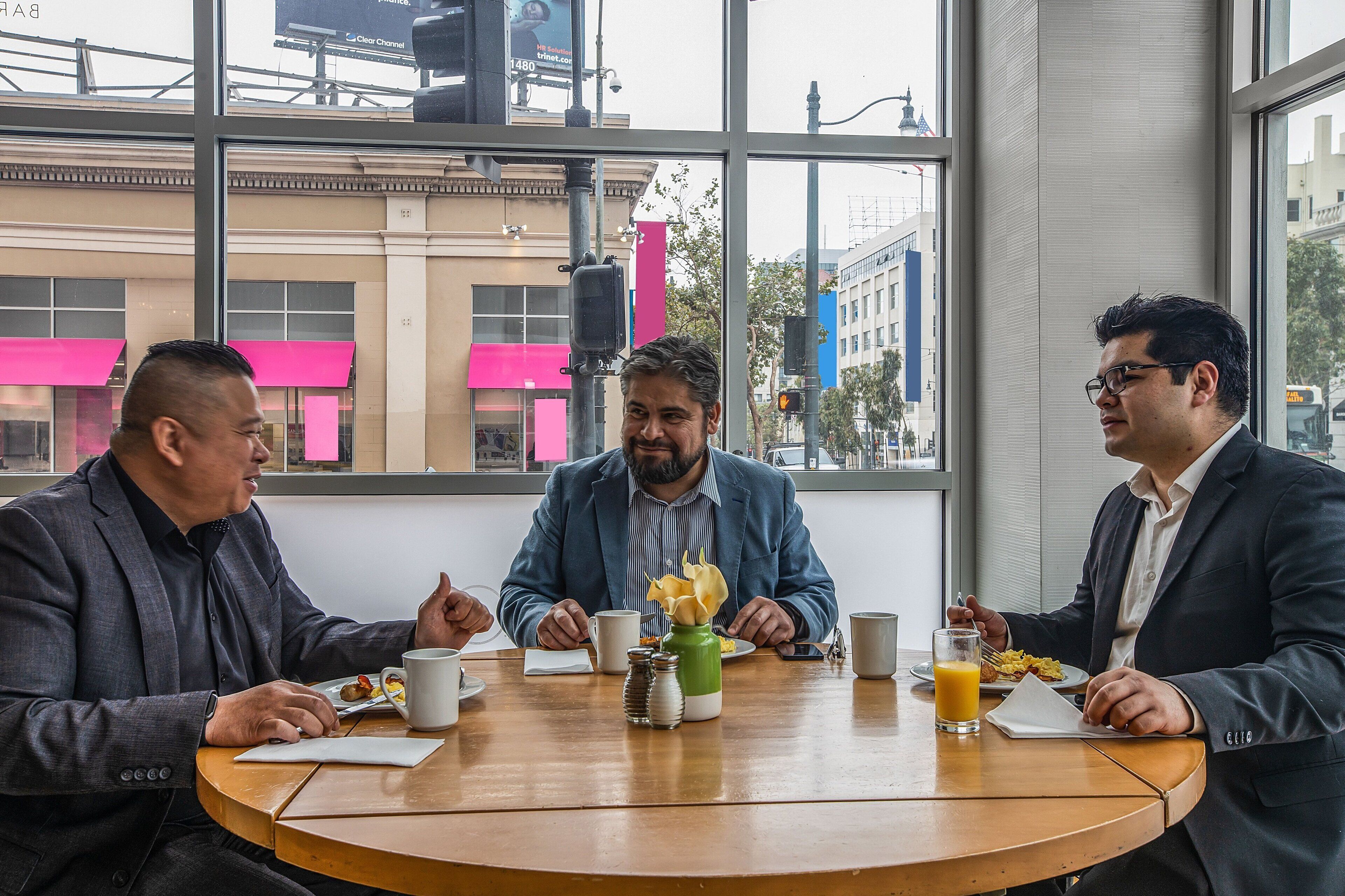 Three men sitting at a round wooden table in a modern indoor setting with large windows.