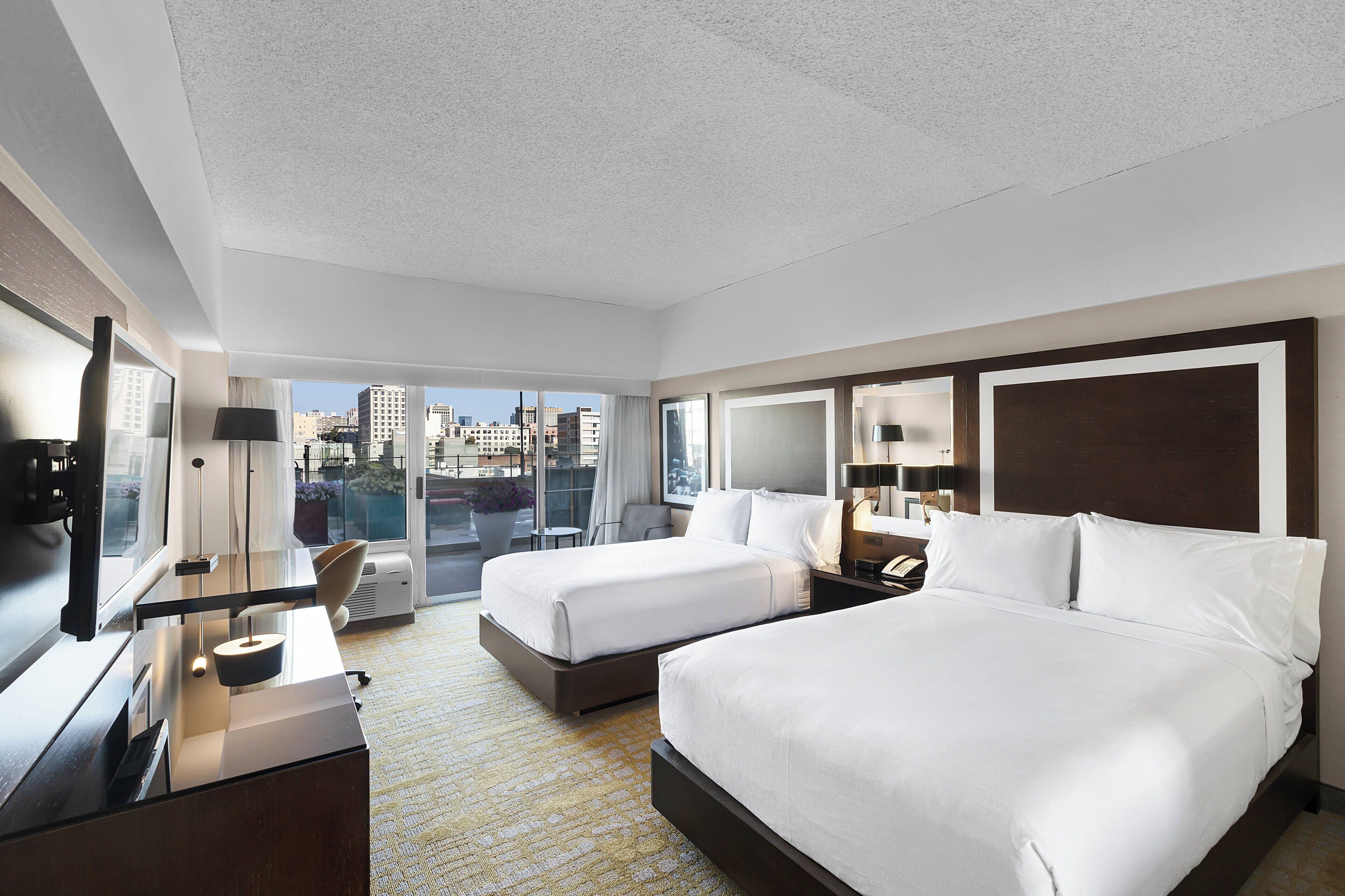 Quarto de hotel com duas camas, uma mesa, uma cadeira, uma televisão e uma janela grande com vista para a cidade.