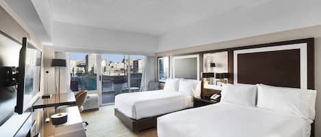 A hotel room with two beds, a desk, a chair, a TV, and a large window with a city view.