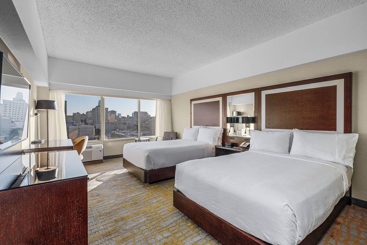 A hotel room with two beds, a desk, a chair, and a large window offering a city view.