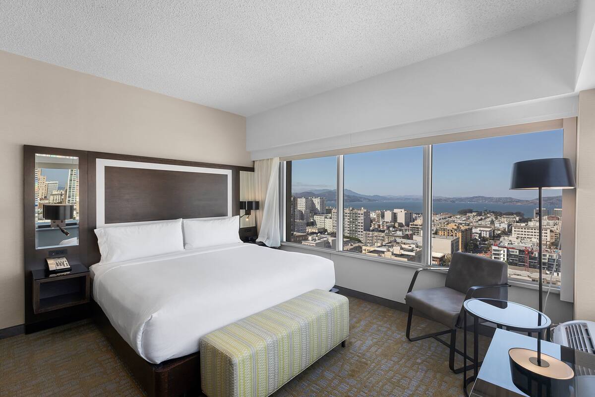 A hotel room with a large bed, a small table, a chair, and a view of the city through large windows.