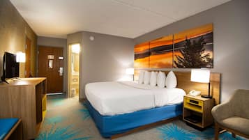 A hotel room with a bed, a desk, a chair, a nightstand with a lamp, and a painting on the wall.