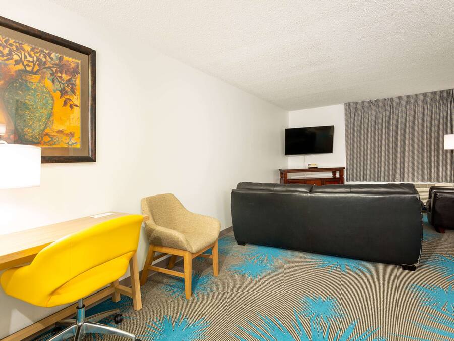 Quarto de hotel com escrivaninha, uma cadeira amarela, sofá preto e televisão, além de carpete estampado.