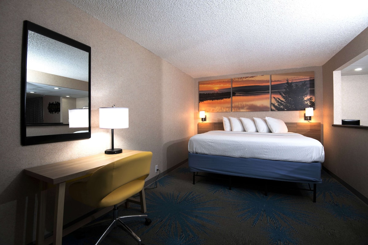 A hotel room with a bed, a desk, a chair, a large mirror, and a wall-mounted lamp.