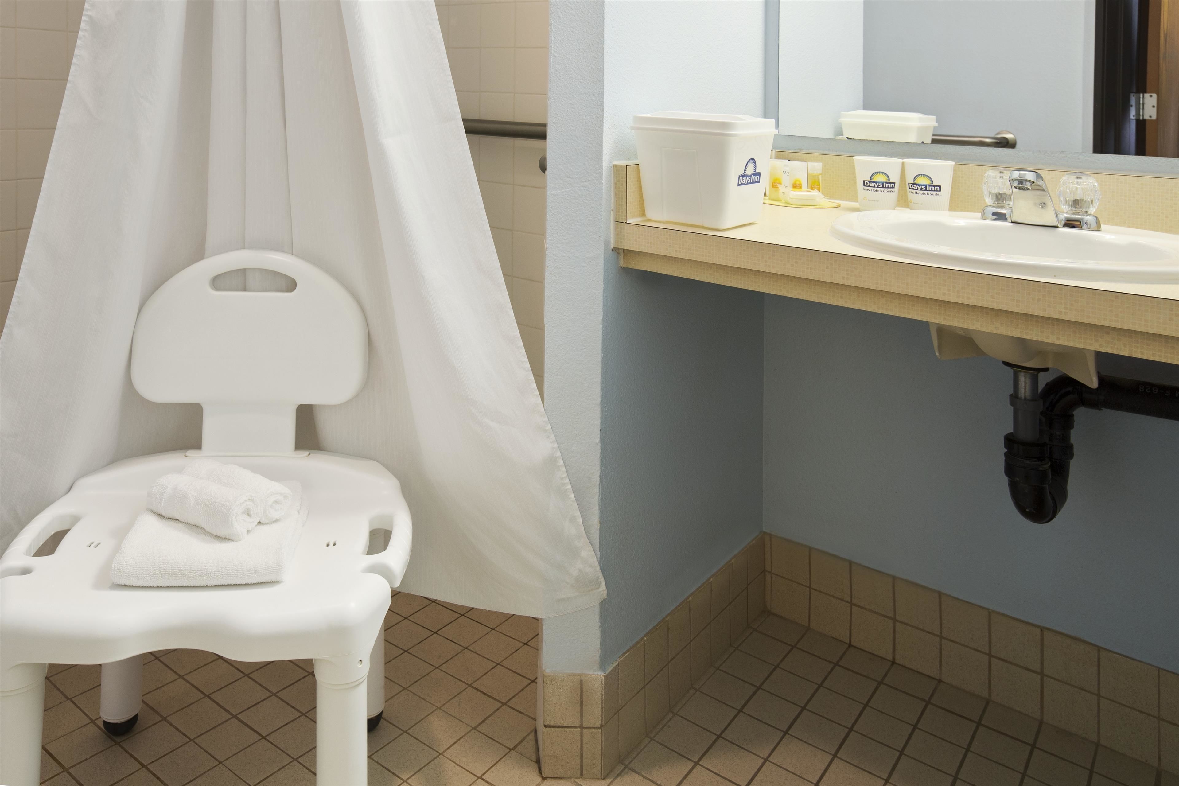 A bathroom with a white shower chair, a white towel, a sink with toiletries, and a mirror.